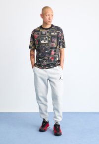 Grafisk t-shirt med blandede billeder, sort bund med farverige mønstre. Grå sweatpants med logo, sorte og røde sneakers med accenter.