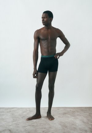 Ψηλός, λεπτός άνδρας, όρθιος ξυπόλητος πάνω σε χαλί, φοράει μαύρα μποξεράκια με πράσινη μέση, κοιτάζει αριστερά σε ένα απλό φωτεινό φόντο.