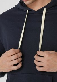 Navyblauer Hoodie mit cremefarbenen Kordeln, ausgestattet mit einer Vordertasche. Der Stoff wirkt weich, hat eine glatte Textur und eine lockere Passform.