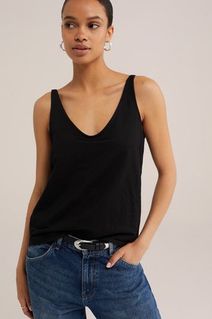 Femme portant un haut noir sans manches rentré dans un jean bleu avec une ceinture noire, debout avec une main dans la poche sur un fond neutre.