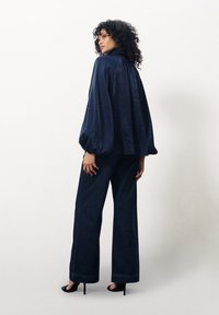 Blouse bleu marine à manches longues avec des manches volumineuses, associée à un jean bleu marine à jambes larges et des chaussures à talons hauts noires. Texture de tissu lisse.