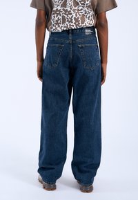 Jeans in denim blu scuro con una vestibilità ampia, dotati di tasche posteriori e un design a gamba dritta, abbinati a una camicia marrone a trama.
