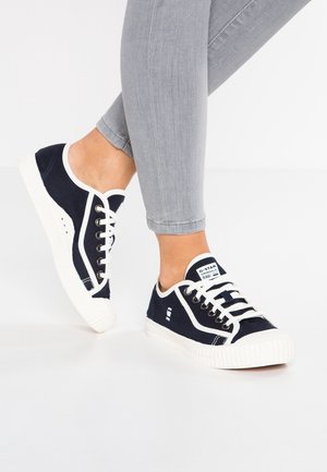 Person trägt graue Slim-Fit-Hose und marineblaue Canvas-Sneaker mit weißen Sohlen und Schnürsenkeln, steht auf weißem Boden.