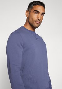 Sweat-shirt bleu à manches longues en tissu texturé, avec un col rond et une coupe droite. Couture d'accent autour de l'encolure.