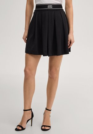 Woman wearing black pleated skirt with JOOP! JEANS waistband and black ankle-strap high heels, standing against plain background.