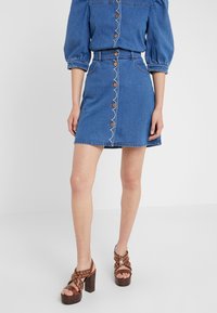 Denimová mini sukně s předními knoflíky a vykrojeným designem. Vyznačuje se světle modrou barvou, kapsami a kontrastním šitím. Součástí jsou hnědé sandály na podpatku.