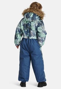 Vaikas apsirengęs mėlynai-žaliu gėlių rašto žiemos sniegui skirtu kostiumu su pūkuotu kapučinu, stovi atsisukęs nugara prieš baltą fono.