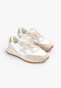 Zapatillas blancas y beige con cordones rosas, detalles dorados y un logo de calavera en el lateral. Materiales con textura y suela de goma.