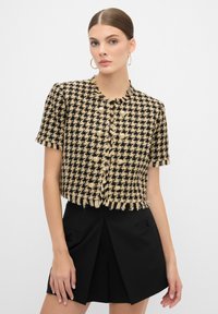 Svart och beige tweedjacka med korta ärmar, fransdetaljer och runt, ljusfärgade knappar, ihopparad med svarta högmidjade shorts.