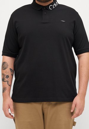 Muž nosící černé polo tričko s krátkým rukávem s bílým logem Calvin Klein na límci a béžové kalhoty, viditelný tetování na paži.