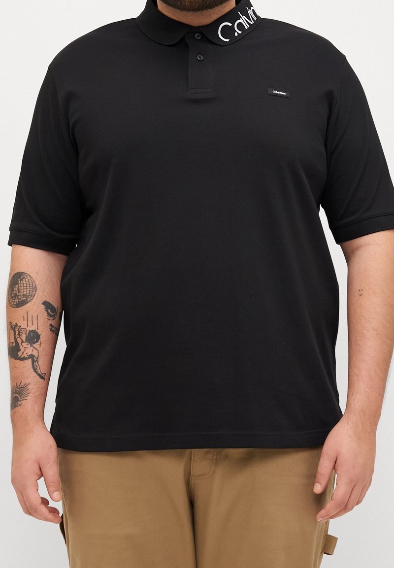 Homme portant un polo noir à manches courtes avec un logo blanc Calvin Klein sur le col et un pantalon beige, tatouage visible sur le bras.