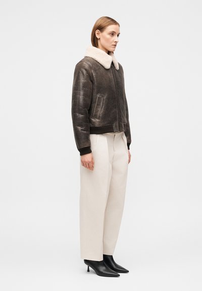 Καφέ μπουφάν bomber από shearling με κρεμ γιακά, κλείσιμο με φερμουάρ και ριμπ μανσέτες, συνδυασμένο με κρεμ παντελόνι φαρδιάς γραμμής και μαύρες μυτερές γόβες.