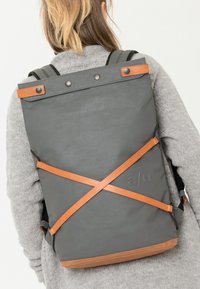 Grauer Rucksack mit tanfarbenen Lederakzenten, strukturiertem Finish, oberen Druckknöpfen und überkreuzten Riemen, getragen über einem grauen Strickpullover.