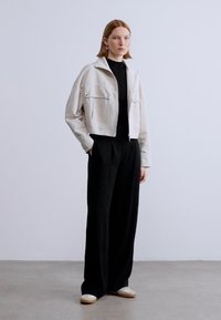 Λευκό κοντό τζάκετ με δύο μπροστινές τσέπες, φορεμένο πάνω από μαύρο πουλόβερ, συνδυασμένο με άνετο μαύρο παντελόνι και ανοιχτόχρωμα παπούτσια.