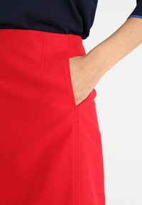 Jupe rouge présentant une texture lisse, une poche latérale et des coutures nettes. Le design est ajusté avec une silhouette droite.