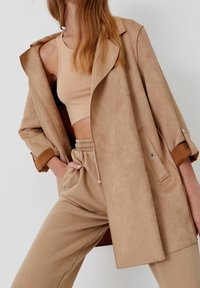 Beige suede jacket with a relaxed fit, brown cuff accents, and side pockets, paired with coordinating tan cropped top and pants.