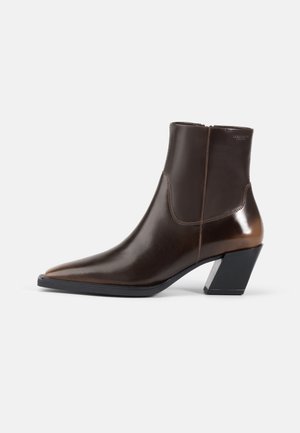 Bottine en cuir brun avec un bout pointu et un talon noir géométrique. Texture lisse avec un panneau contrastant et une fermeture éclair.
