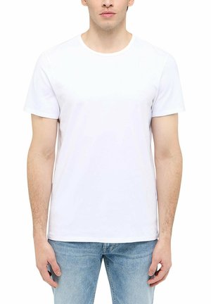 Mann trägt ein schlichtes weißes T-Shirt mit kurzen Ärmeln und hellblaue Jeans, steht mit entspannten Armen seitlich am Körper vor weißem Hintergrund.