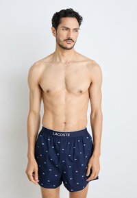 Navy boxers with blue embroidered logos, button fly, and elastic waistband featuring "LACOSTE" branding. Smooth fabric, casual design.