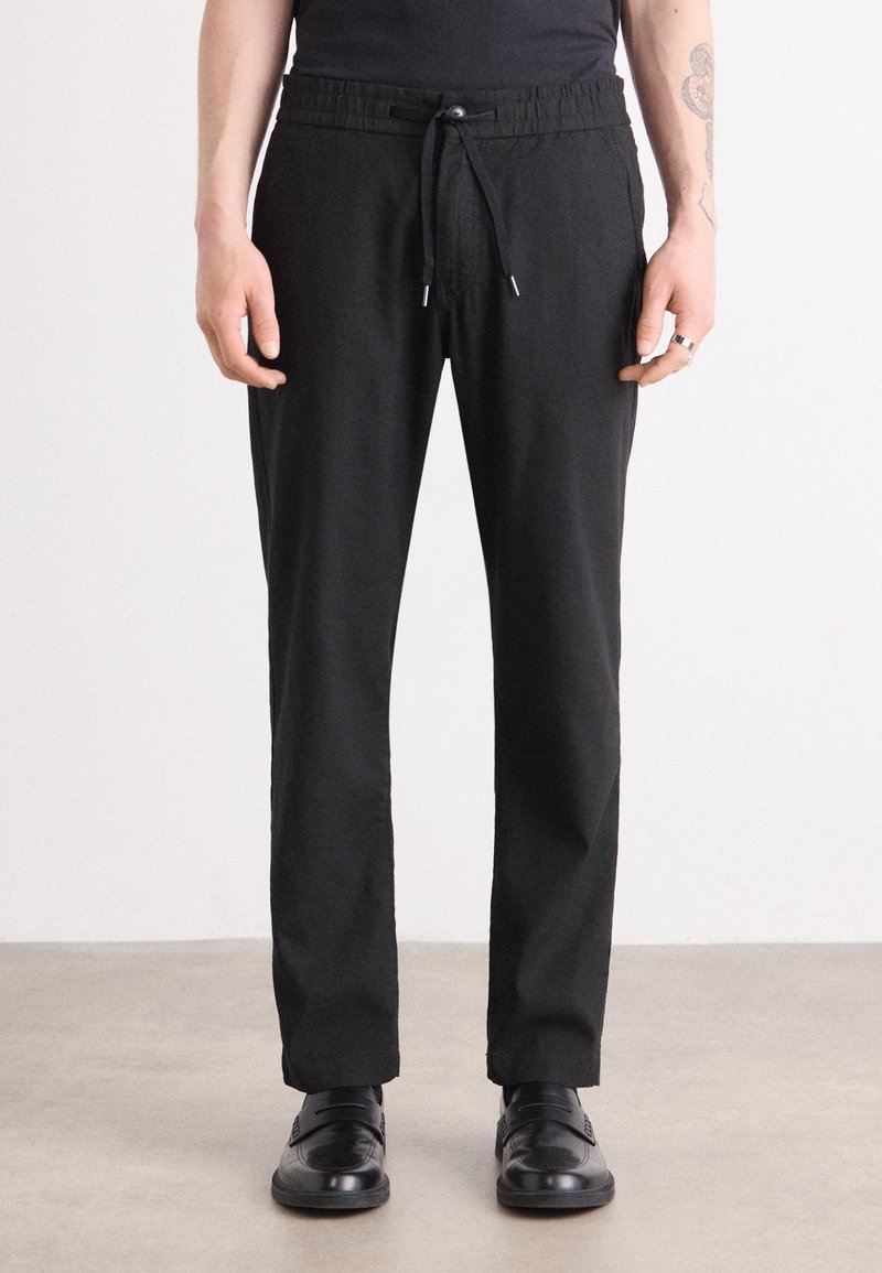 Homme portant un pantalon noir à cordon, chaussures en cuir noires, chemise noire, debout sur un sol gris contre un mur blanc.