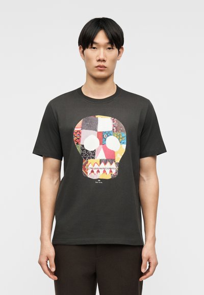 Jeune homme portant un t-shirt noir avec un grand motif de crâne en patchwork coloré sur le devant, debout devant un fond gris uni.