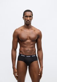 Čierne krátke spodné prádlo s elastickým pásom a logom "HUGO". Hladká textúra, priliehavý dizajn a minimalistický štýl. Model stojí vzpriamene.