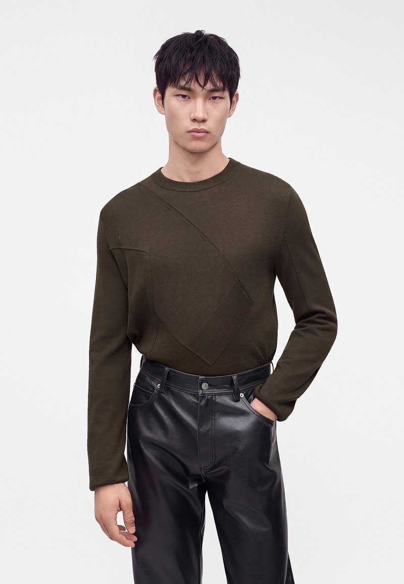Jeune homme portant un pull à manches longues vert olive avec des détails diagonaux texturés et un pantalon en cuir noir, debout devant un fond uni.