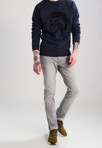 Felpa blu navy con stampa grafica, jeans grigi e sneakers verde oliva. Tatuaggi notevoli sulle braccia, design casual, vestibilità moderna.