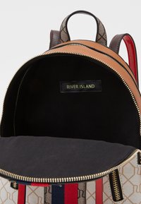 Tan och mönstrad tygryggsäck med svart inredning, dragkedja i guld, och en logotags invändigt. Har röda och blå detaljer.