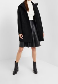 Svart ullblandad kappa med stor krage och framfickor, parat med en svart läderskjorta och ankelskor. Slät textur och minimalistisk design.