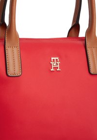 Κόκκινη τσάντα χειρός από νάιλον με καφέ δερμάτινες λαβές, με χρυσό λογότυπο ως διακοσμητικό στοιχείο. Ομαλή υφή και δομημένο σχήμα με αντίθετες λεπτομέρειες.