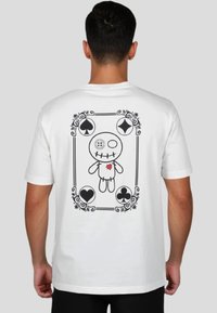 Camiseta blanca de algodón con un gráfico negro de una muñeca con un detalle de corazón y símbolos de cartas enmarcados en bordes decorativos en la parte trasera.
