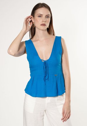 Haut peplum en lin bleu avec un décolleté en V profond et un détail noué à l'avant. Doté de larges bretelles et d'une étiquette métallique sur le côté. Associé à un pantalon blanc.