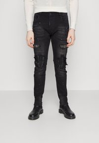 Vaqueros de mezclilla negros con parches desgastados, decorados con lentejuelas negras. Diseño ajustado con un ligero desvanecimiento y detalles de hardware mínimos.