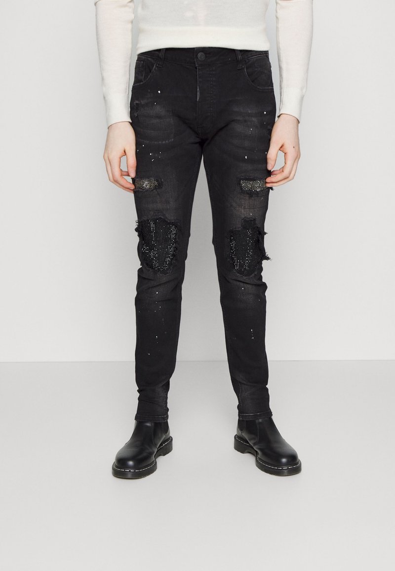 Vaqueros de mezclilla negros con parches desgastados, decorados con lentejuelas negras. Diseño ajustado con un ligero desvanecimiento y detalles de hardware mínimos.
