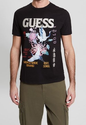 Czarny t-shirt z krótkim rękawem z logo "GUESS", kwiatowym motywem i grafiką żurawia, napisem "WORLD WIDE BRAND" i "EST. 1981" w kolorze żółtym.