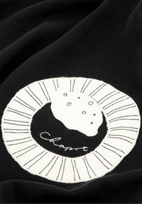 Zwarte stof met een cirkelvormige witte geborduurde patch met een gestructureerd ontwerp van een roomgevuld gebakje, omringd door lineaire accenten en tekst.