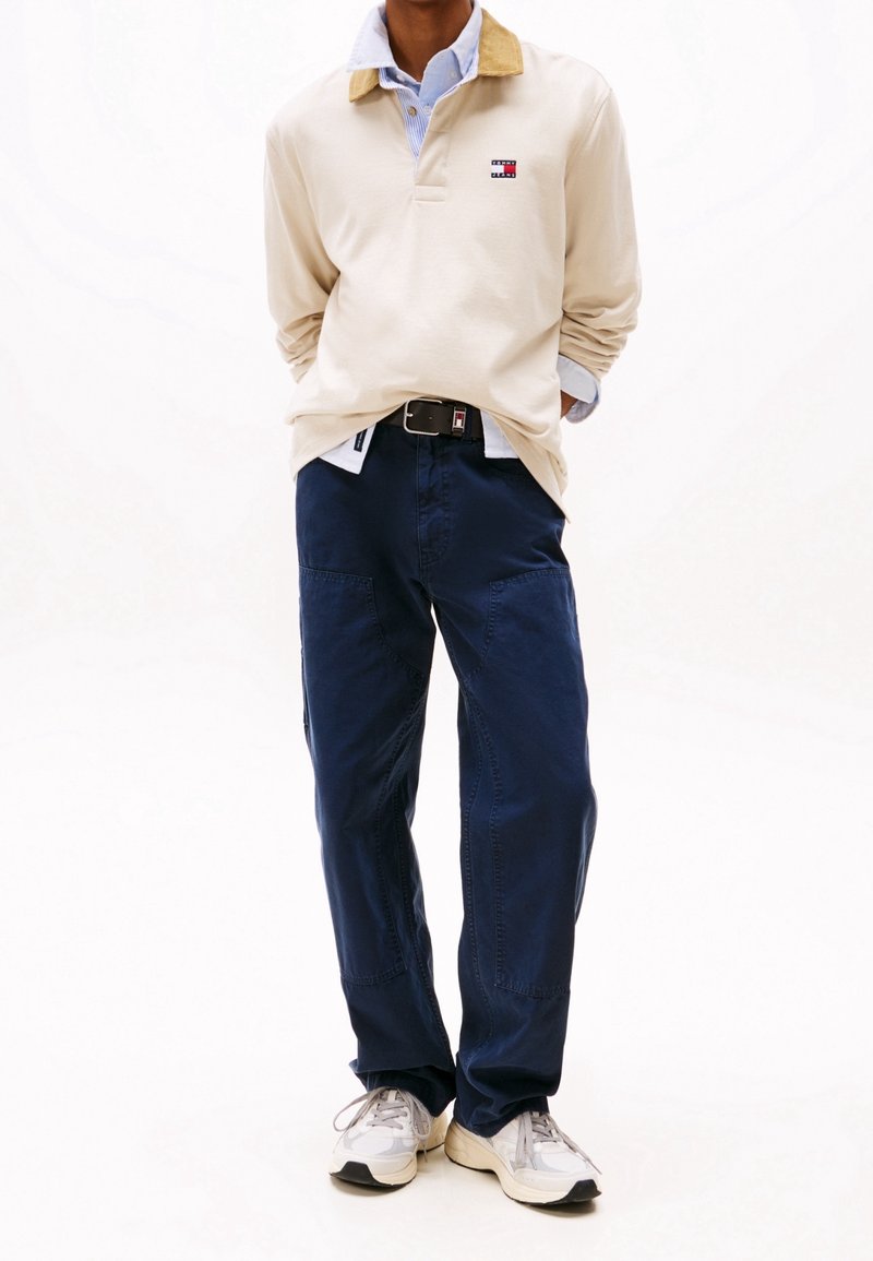 Polo beige à manches longues avec un col en velours côtelé, pantalon cargo bleu marine à coupe décontractée et baskets grises ; présente un petit logo sur la poitrine.