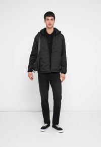 Svart boblevest med hette over en svart skjorte, kombinert med mørke jeans og svarte joggesko. Har en glatt tekstur og glidelåslukking.