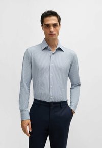 Chemise boutonnée à manches longues, bleu clair avec un motif fin à rayures, associée à un pantalon sombre et ajusté. Tissu lisse et style près du corps.