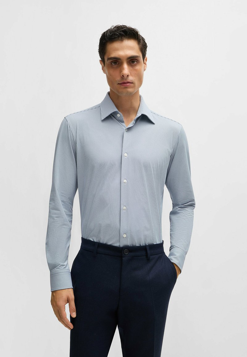 Chemise boutonnée à manches longues, bleu clair avec un motif fin à rayures, associée à un pantalon sombre et ajusté. Tissu lisse et style près du corps.