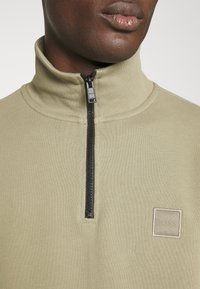 Světlá olivově zelená mikina na zip s vysokým límcem, černým zipem a čtvercovou nášivkou s logem na hrudi. Hladká textura látky.