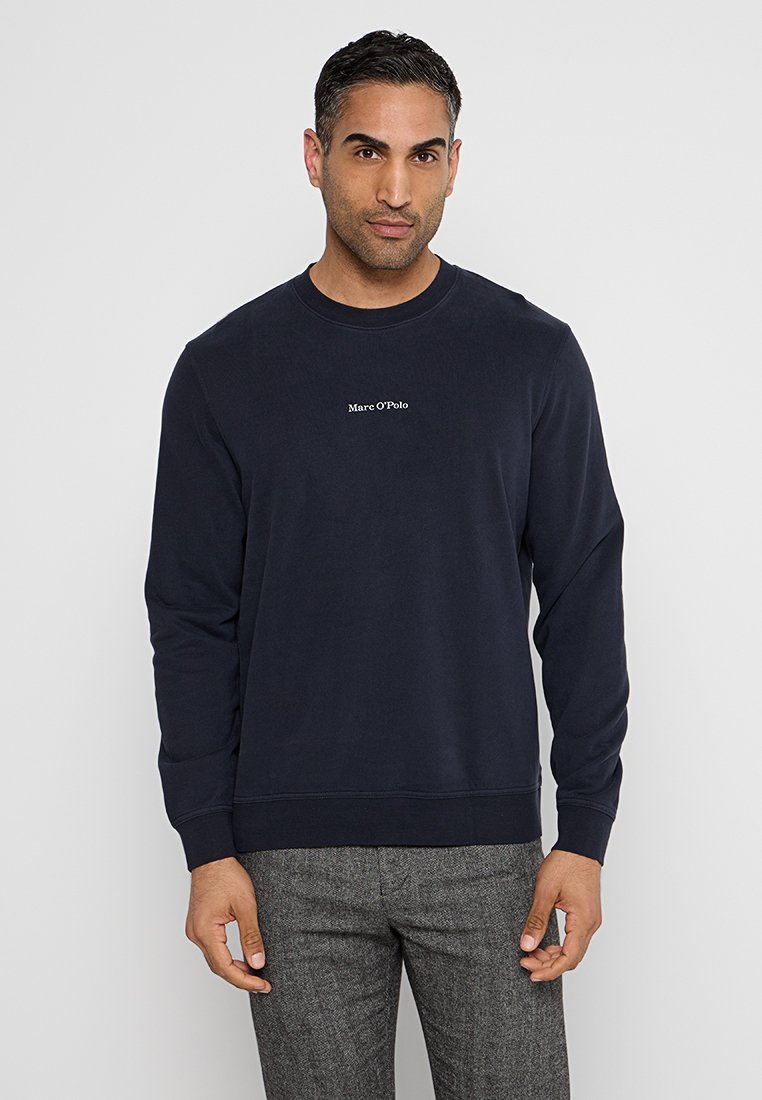 Homme portant un sweat-shirt bleu marine de Marc O'Polo et un pantalon gris texturé, se tenant contre un fond gris clair uni.