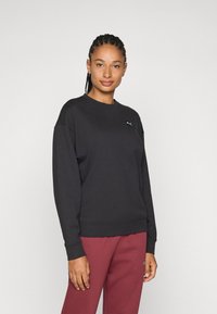 Sudadera negra con cuello redondo y diseño de hombros caídos, hecha de un tejido suave. Presenta un pequeño logo en el pecho, combinada con pantalones color burdeos.