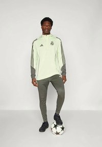 Giacca sportiva verde chiaro con zip, dettagli neri, maniche grigie e logo. Pantaloni sportivi grigi e sneaker neri. Tieni in mano un pallone da calcio.