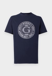 Marineblå bomulds-t-shirt med et cirkulært hvidt grafik, der inkluderer tekst som "Guess" og "Los Angeles" med fede designelementer.