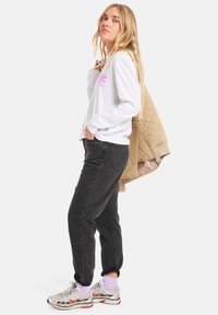 White long-sleeve shirt with pink logo, paired with black jeans and silver sneakers. Holding a tan quilted jacket.