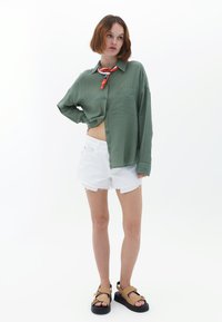 Camicia verde a maniche lunghe con taschino, abbinata a pantaloni corti bianchi con bordi sfrangiati. Il modello indossa sandali marroni con dettagli in cinghia e una sciarpa multicolore.