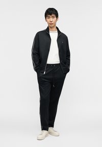 Μαύρο μπουφάν με φερμουάρ και λευκές ρίγες, φορεμένο πάνω από λευκό πουκάμισο. Μαύρο παντελόνι φόρμας με κορδόνι, σε συνδυασμό με κρεμ παπούτσια αθλητικά.