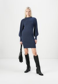 Marineblauwe geribbelde gebreide jurk met lange pofmouwen, gecombineerd met zwart leren laarzen tot de knie met blokhak en een zwarte handtas.